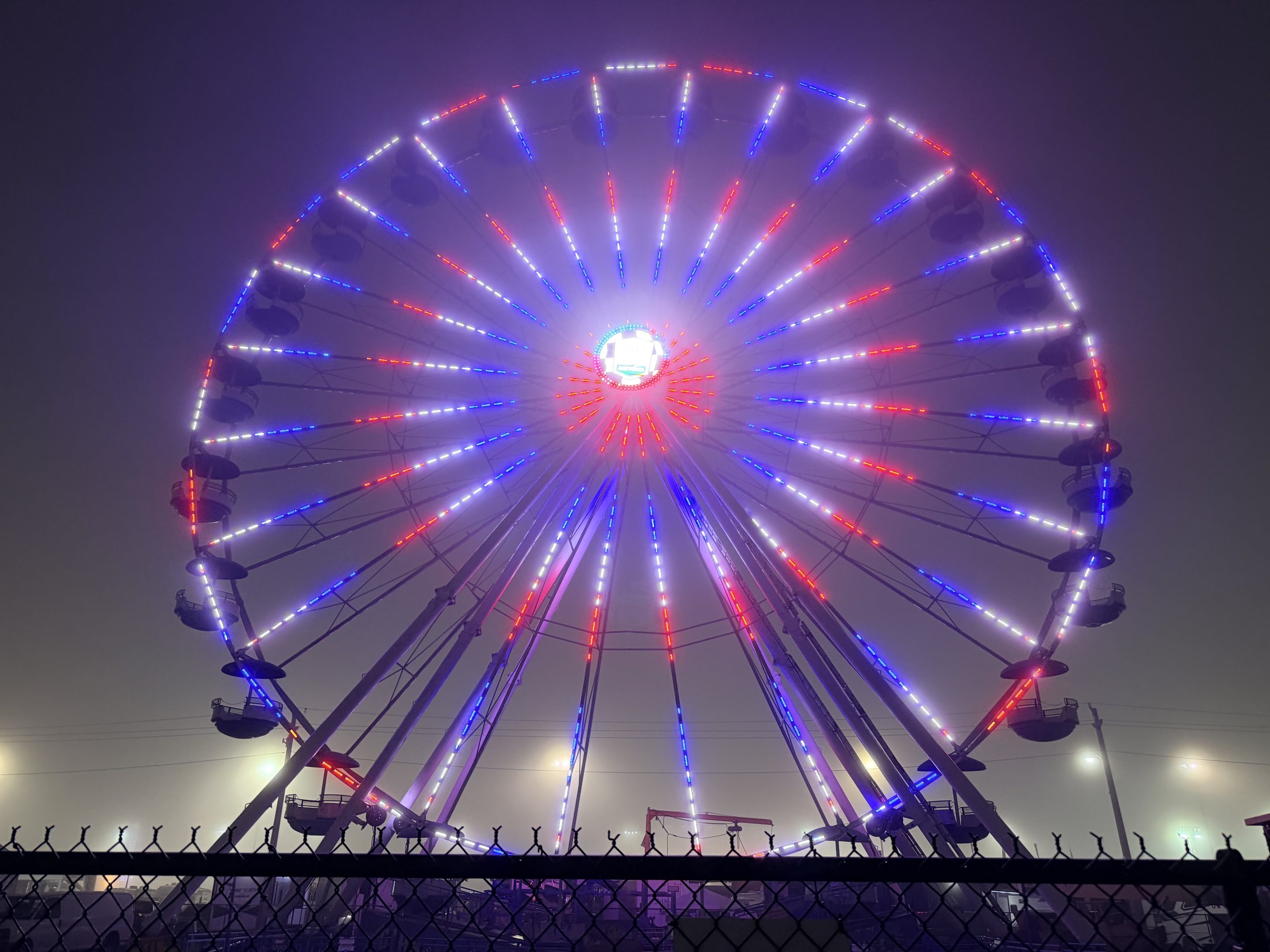 The ferris wheel at Daytona International Speedway lit in red, white, and blue and backlit by the track lighting, all immersed in thick fog at night, with the top of a chainlink fence in the foreground.